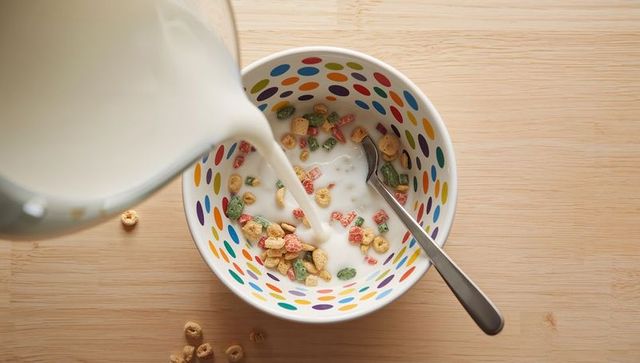 Pouring milk into colorful polka-dot bowl with fruity oat cereal, overhead breakfast on wood table