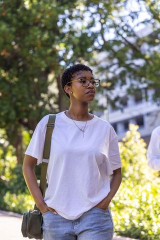 Woman of african descent standing on sunlit walkway wearing glasses while carrying bag