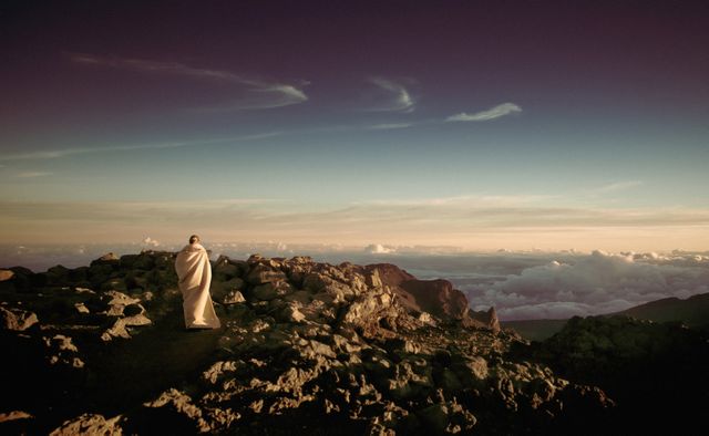 This image depicts a solitary figure wrapped in a blanket standing on a rocky summit at sunrise. The expansive sky and clouds underscore peaceful and reflective themes, evoking a sense of solitude and connection with nature. Ideal for use in articles or presentations on hiking, travel, nature retreats, meditation, and the majesty of alpine landscapes.