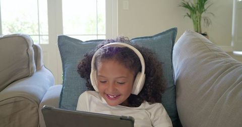 Smiling child wearing headphones watching tablet on cozy gray sofa with blue pillow