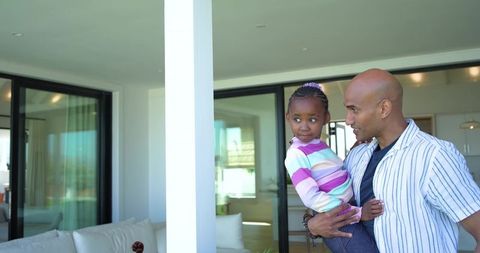 Father holding african american daughter near glass doors in modern home, family bonding
