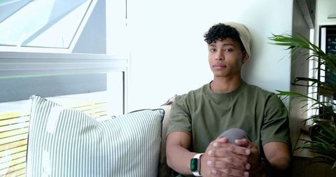 Young man sitting on sunlit window seat wearing beanie and smartwatch in minimalist home