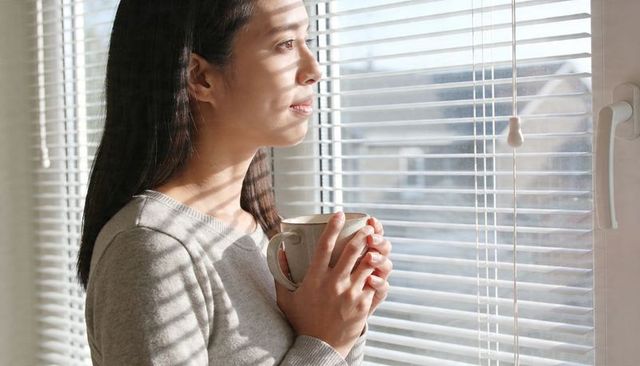 Asian woman holding mug looking through blinds in sunlit room for morning reflection