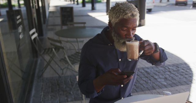 African american senior man using smartphone while drinking coffee sitting outdoors at cafe. active senior lifestyle living concept