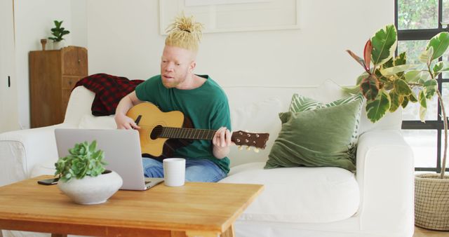 Man playing acoustic guitar while following along on laptop screen. Sitting on white sofa in bright and modern living room with green plants and wooden table. Ideal for use in articles about online music lessons, hobbies at home, or cozy home settings.