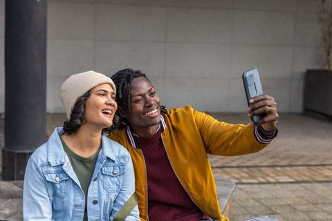 Young couple taking selfie on urban plaza bench with carry-on luggage, casual autumn style