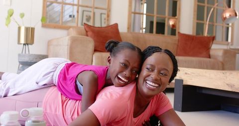 African american mother and daughter laughing on living room floor wearing pink tops cozy bonding