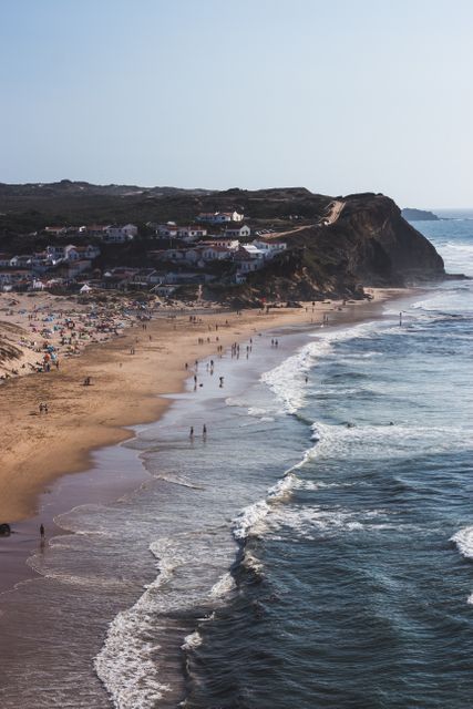 Aerial photograph capturing a crowded beach with people relaxing and enjoying in summer. The waves crash onto the shore, next to a scenic cliff with houses visible in the backdrop. Perfect for travel blogs, tourism promotions, or websites featuring tropical vacation destinations.