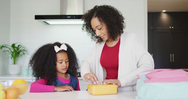 Mother with curly hair is preparing a lunchbox for her young daughter in a modern kitchen. The daughter is standing by attentively. The scene shows family routines, parental care, and everyday home life. Ideal for advertisements, parenting blogs, family lifestyle magazines, and educational materials.