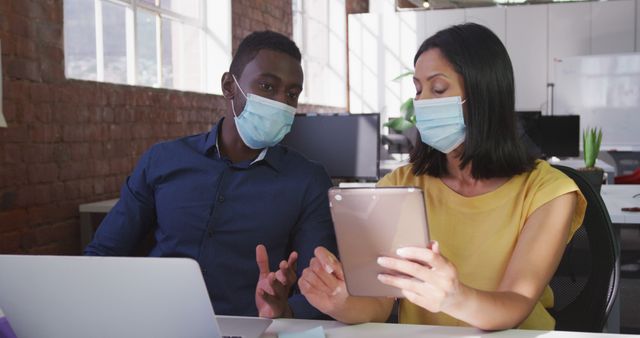 Two colleagues discussing project using tablet while wearing face masks in a modern office. Ideal for illustrating workplace safety measures, remote communication, teamwork, and technological collaboration during the pandemic. Represents adherence to health protocols and adaptability in business settings.