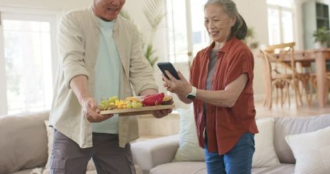 Senior asian couple sharing vibrant fruit tray while woman taking smartphone photo