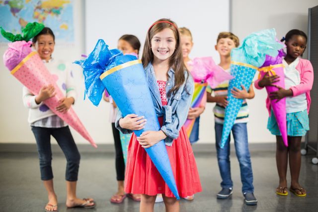 Children standing in a classroom holding colorful gift cones, smiling and celebrating. Perfect for educational materials, school event promotions, and advertisements focusing on childhood joy and diversity.