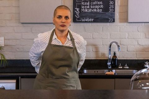Barista standing behind coffee counter wearing green apron and cropped hair in modern cafe