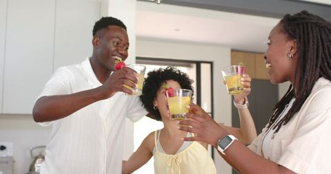 African american friends cheering with lemonade and strawberry garnish in bright kitchen