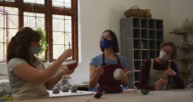Three people engaging in pottery work in an art studio, all of them are painting ceramic items while wearing protective masks. Can be used for themes relating to creativity, teamwork, art and craft classes, small business, or artistic hobbies.