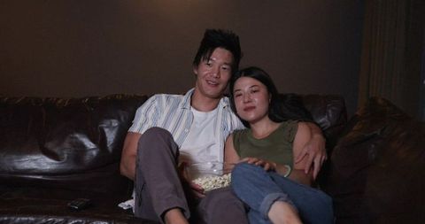 Asian couple cuddling on dark leather sofa sharing popcorn while watching movie at home