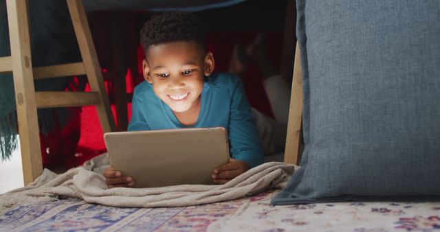 Smiling boy lying under blanket fort using digital tablet. This image exemplifies childhood creativity and indoor playtime. Perfect for advertisements related to educational apps, children’s digital entertainment, technology for kids, or home learning environments.