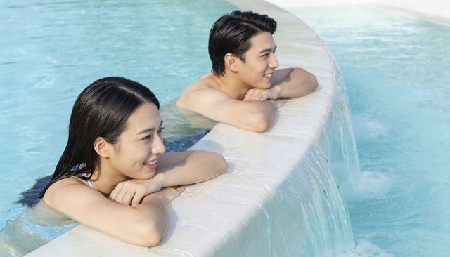 Young couple relaxing at curved pool edge with cascading water, resort leisure getaway