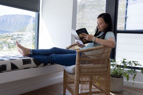 Mature asian woman reading paperback in rattan chair by sunlit window with mountain view