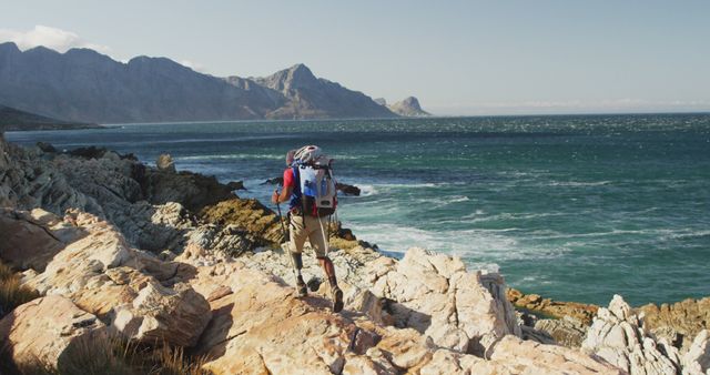 Person hiking along a rocky coast with mountains in the background, ideal for travel blogs, outdoor adventure promotions, hiking gear advertisements, and nature-themed websites. Floating sky above blue-green ocean waves hints to new adventures and untamed natural beauty.