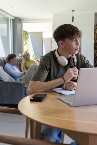 Teen studying at home with headphones and laptop while parents relaxing in living room