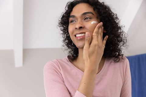 Non-binary person applying face moisturizer in bright bathroom, smiling during self-care routine
