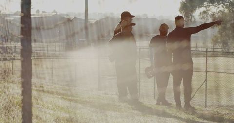 Backlit team of men conferring and pointing on slope with chain-link fence at golden hour