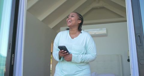 Laughing african american woman in mint loungewear holding remote on sunlit bedroom balcony