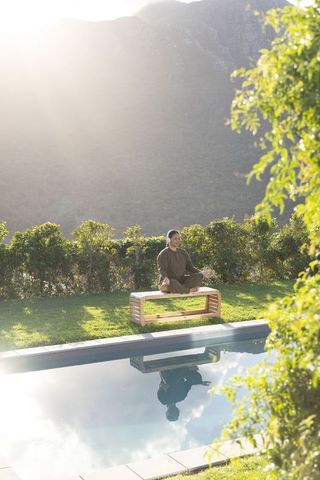 Sunlit asian woman meditating on wooden bench by pool with mountain reflection