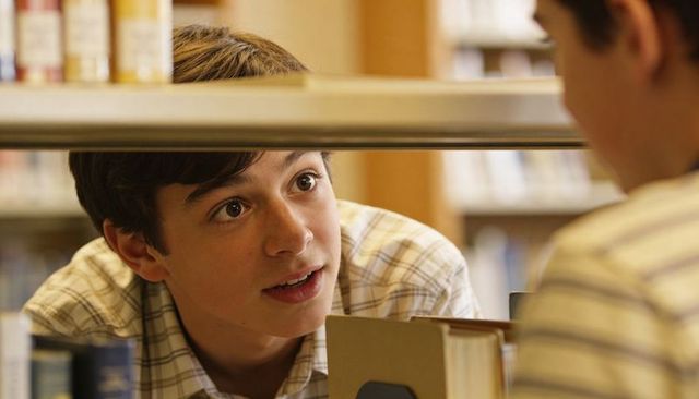 Curious teenage boy peering under library shelf while studying, plaid shirt student moment