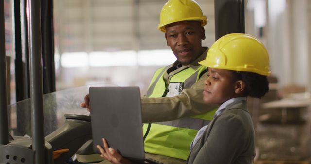 Two warehouse workers wearing safety helmets and vests are engaged in working on a laptop. The male worker sits on a forklift while the female employee stands, holding the laptop. They are in a modern warehouse environment, likely discussing or managing inventory. This image can be used for themes related to industrial work, warehouse logistics, teamwork, safety protocol, and technology in manual labor settings.