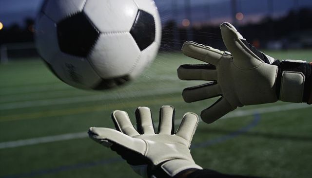 Goalkeeper reaching and catching soccer ball on wet turf at dusk under floodlights