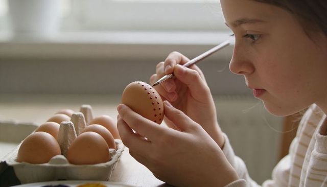 Child decorating brown egg with dotted paintbrush at home table