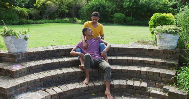 Smiling biracial gay male couple sitting on steps hugging in garden. staying at home in isolation during quarantine lockdown.