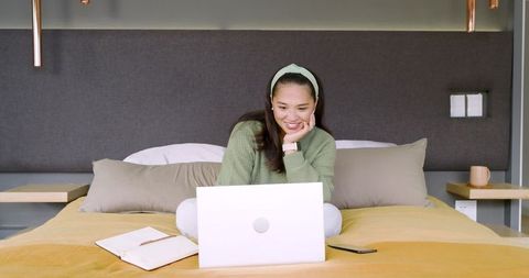 Asian woman enthusiastically engaging with laptop in modern bed setting