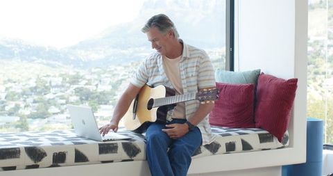 Mature man playing acoustic guitar on window seat while using laptop with mountain view