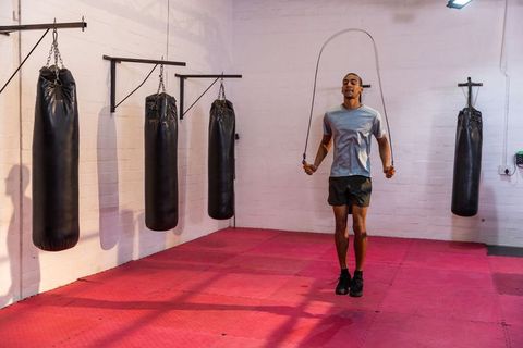 African american athlete jumping rope in boxing gym with heavy bags and red mats