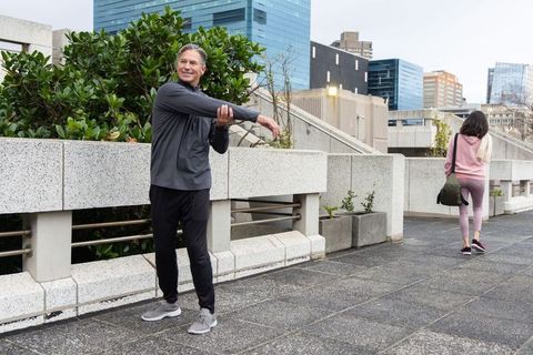 Middle-aged man stretching on urban terrace while woman walking past modern city skyline