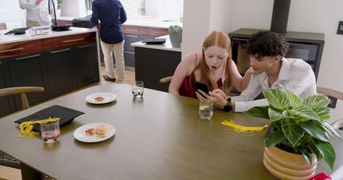 Couple leaning over smartphone beside graduation cap on dining table, casual celebration
