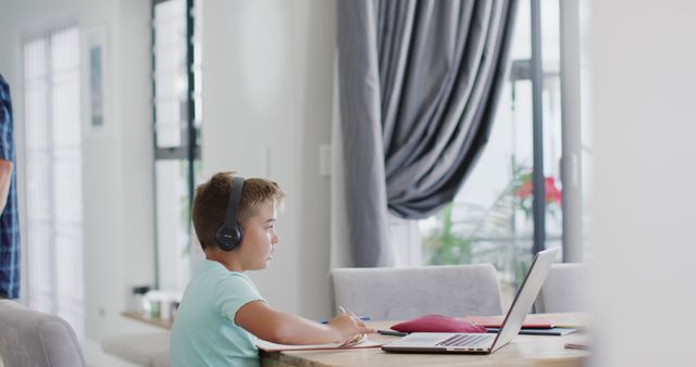 Young boy engaging in online education from home, using laptop and headphones while sitting at table. Ideal for illustrating concepts of e-learning, homeschooling, remote education, and children's use of technology. Suitable for educational websites, online teaching resources, and parenting blogs.
