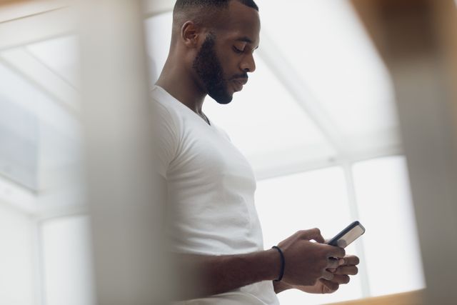This image shows an African-American man using a smartphone indoors, perfect for illustrating concepts related to technology, communication, and modern lifestyle. Suitable for use in articles, blogs, and advertisements focusing on mobile technology, connectivity, and everyday life.