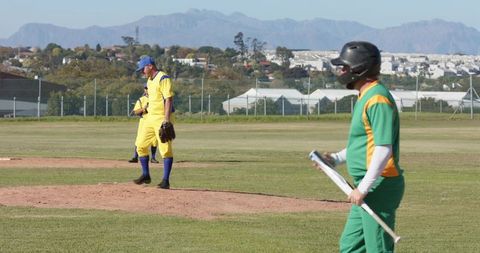Mid-adult batter holding metallic bat while pitcher standing on mound in suburban baseball game