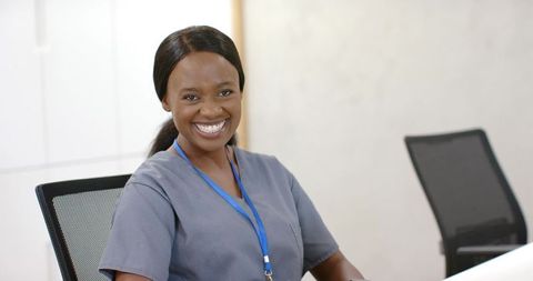 Smiling black nurse at clinic reception wearing gray scrubs and blue lanyard, welcoming