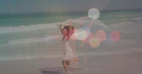 Joyful woman running on sunlit beach holding sheer scarf with bokeh lensflare