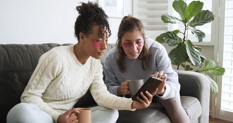 Two women relaxing on couch wearing under-eye gel patches sharing smartphone and coffee