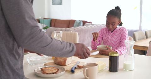 African american father preparing breakfast while daughter stirs cereal at sunlit kitchen