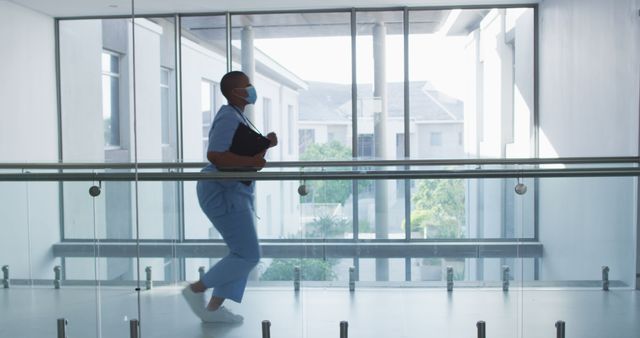 African american female health worker wearing face mask running in the corridor at hospital. medical healthcare during coronavirus covid 19 pandemic concept