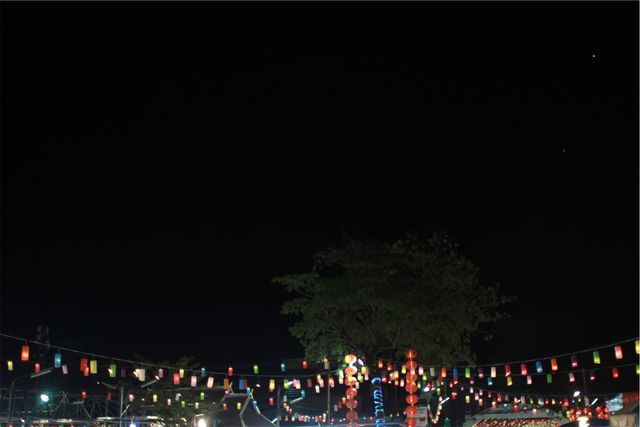 String lights in various colors hanging outdoors during a night festival, creating a festive atmosphere. Ideal for use in articles, advertisements, or social media posts that focus on festivals, outdoor events, celebrations, night parties, or colorful decorations to evoke excitement and joy.