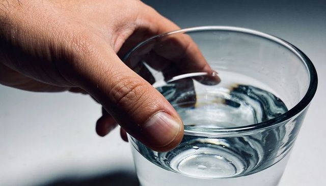 Hand holding glass of water on white countertop, curling around rim, minimalist closeup