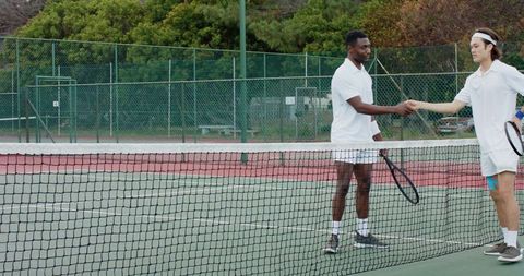 Two male tennis players shaking hands at net after match on outdoor court wearing white